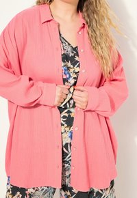 Pink textured button-up shirt with a collar, paired with a dark floral-patterned top underneath. Long sleeves, relaxed fit.