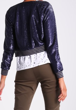 Person wearing a navy blue sequined jacket with striped cuffs over a white star-patterned shirt and brown pants, shown from the back.