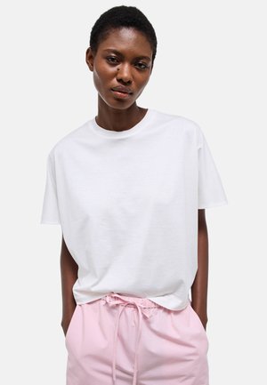 Femme aux cheveux courts portant un t-shirt blanc uni et un pantalon léger rose pâle à cordon, debout les mains dans les poches.