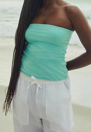 Personne aux longues tresses portant un haut sans bretelles bleu sarcelle et un pantalon blanc à cordon, debout près de la plage avec les vagues de l'océan en arrière-plan.