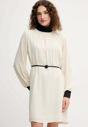 Donna con capelli ricci che indossa un abito crema a maniche lunghe con polsini neri, collo alto nero e sottile cintura nera con fibbia a fiore.