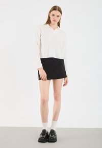 Λευκό πουλόβερ με πλέξη κασμίρ και γιακά, σε συνδυασμό με μαύρη μίνι φούστα. Μαύρα παπούτσια με διακοσμητικές στρογγυλές βίδες και λευκές κάλτσες μέχρι τον αστράγαλο.