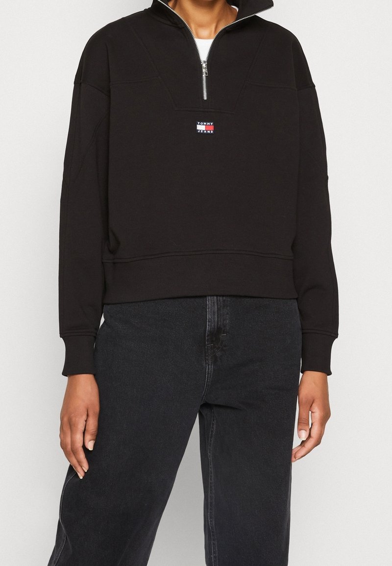Personne portant un pull noir à demi-fermeture éclair avec le logo Tommy Jeans et un jean taille haute foncé sur un fond clair.