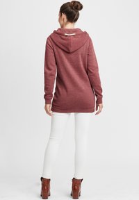 Burgunderfarget hettegenser med teksturert overflate, lange ermer og snor-detaljer i hetten. Kombinert med hvite tettsittende bukser og brune støvler.