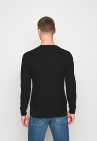 Maglione nero a maniche lunghe con texture a coste, scollatura rotonda e polsini aderenti, abbinato a jeans in denim blu, mostrato di spalle.