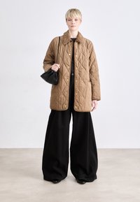 Quilted braune Jacke mit einem kragenartigen Hals, goldenen Druckknöpfen und Taschen, getragen über einem schwarzen Oberteil und weiten Hosen; kombiniert mit einer schwarzen Tasche.