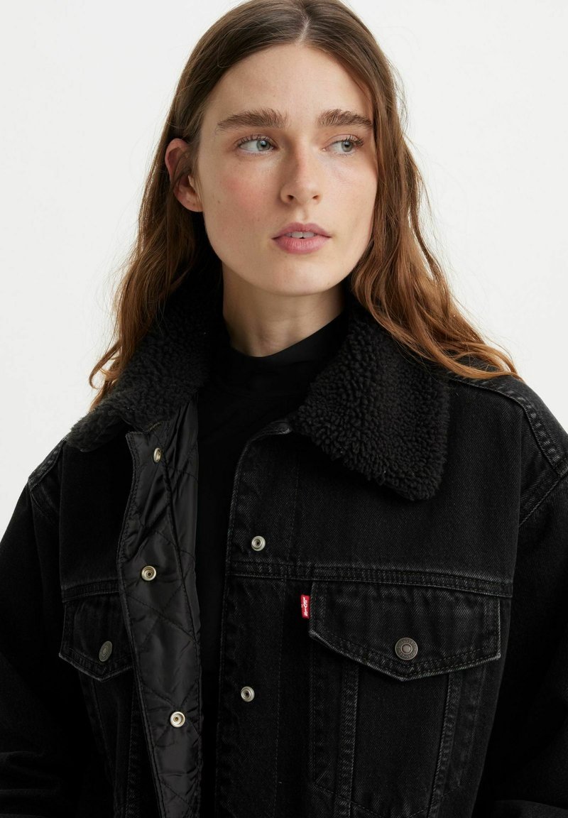 Veste en denim noire avec un col en fourrure noire, boutons-pression et étiquette logo rouge. Comprend un intérieur matelassé et des poches avant.