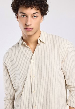 Junger Mann mit lockigem Haar, der ein beiges gestreiftes Hemd mit Knöpfen trägt und neutral geradeaus schaut.