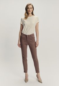Beżowa koszulka z subtelnym kwiatowym wzorem, w zestawieniu z dopasowanymi brązowymi spodniami. Modelka nosi jasnobeżowe szpilki, które prezentują elegancki, minimalistyczny design.