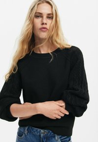 Maglione nero caratterizzato da un design a maglia testurizzata sulle maniche, collo ampio a giro, e polsi a coste, abbinato a jeans in denim blu.