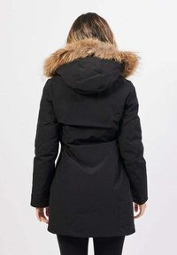 Donna che indossa un cappotto invernale nero con cappuccio bordato in pelliccia marrone, girata di spalle su uno sfondo chiaro e uniforme.