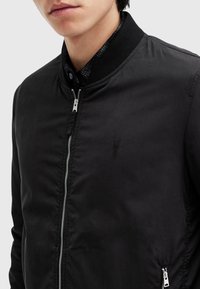 Homme portant une veste bomber noire zippée par-dessus une chemise écossaise foncée, montré des épaules jusqu'au milieu du torse.