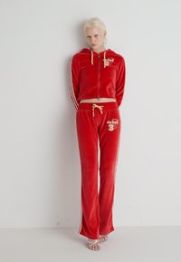 Punainen velour-välikoira, jossa on vetoketjullinen huppari, jossa on valkoiset korostusraidat ja brodeeratut yksityiskohdat, yhdistettynä vastaaviin housuihin ja kevyisiin jalkineisiin.