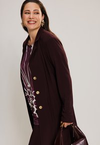 Burgundy long jacket with gold buttons, layered over a patterned purple top. Smooth fabric with a relaxed fit, paired with a dark handbag.