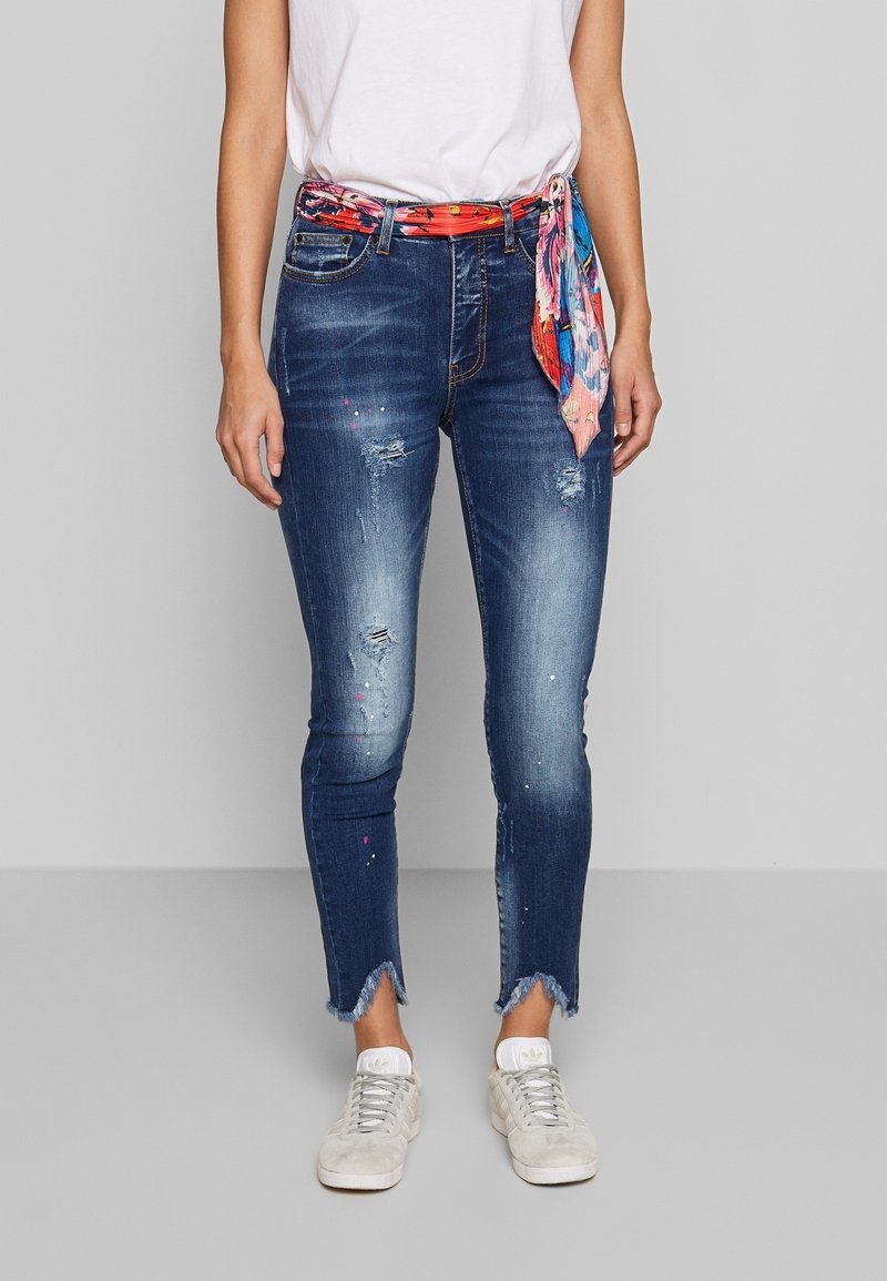 Smala jeans i mörkblå denim med slitningar, fransad nederkant och en färgglad tryckt scarf som är knuten som ett bälte. Kombinerat med vita sneakers.