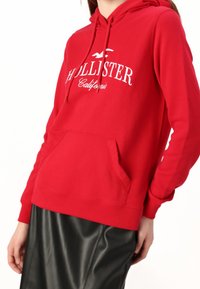 Persona vistiendo una sudadera roja de Hollister California con bolsillo frontal y una falda de cuero negra, de pie contra un fondo blanco.