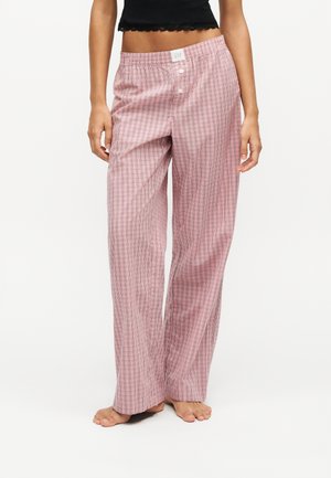 Frau trägt lockere rosa karierte Pyjamahosen und ein schwarzes cropped Top, steht barfuß vor einem einfarbigen Hintergrund.