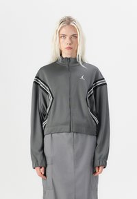 Veste de sport grise, courte, avec un col montant, des rayures blanches sur les manches et un logo sur la poitrine. Le matériau semble lisse avec un léger éclat.