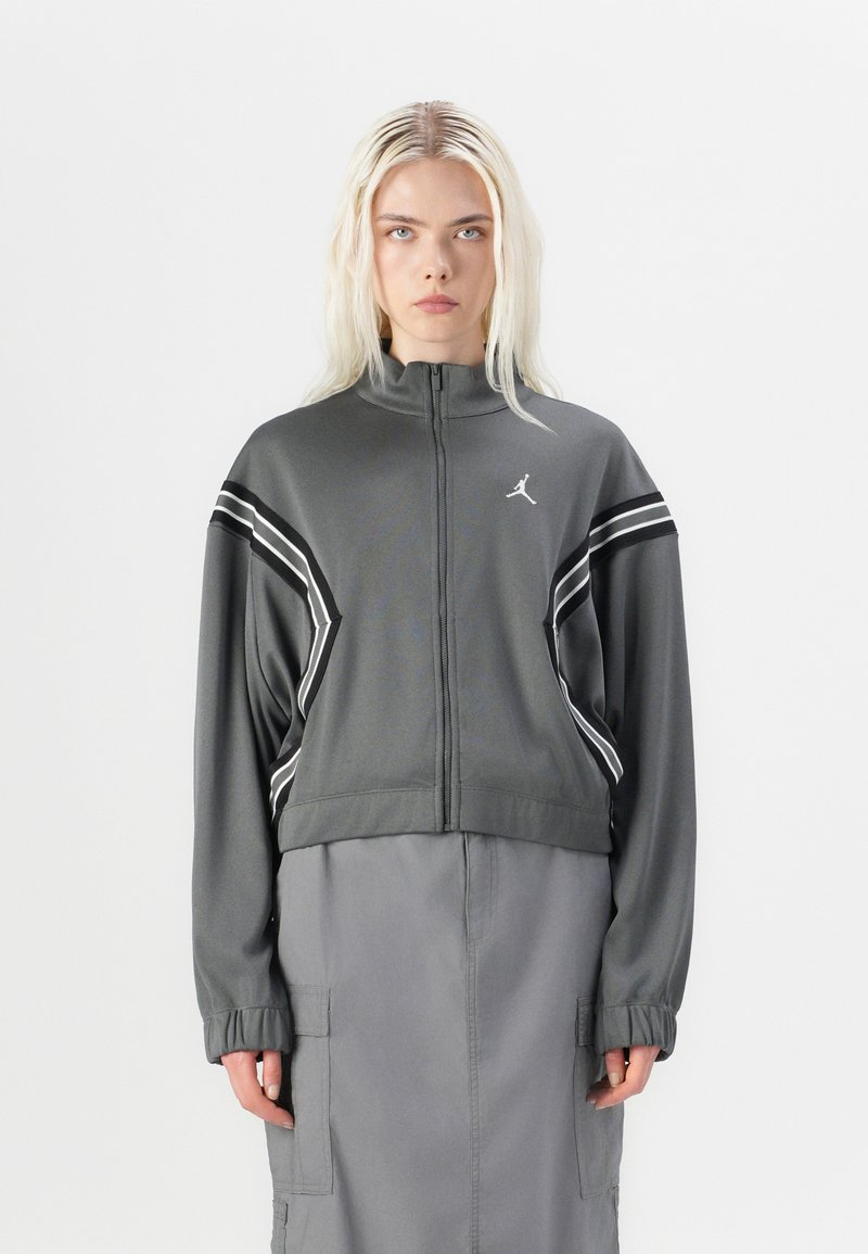 Veste de sport grise, courte, avec un col montant, des rayures blanches sur les manches et un logo sur la poitrine. Le matériau semble lisse avec un léger éclat.