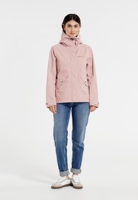 Lichtroze waterdichte jas met capuchon, twee voorzakken en een ritsluiting. Draag het over een blauwe jeans en witte sneakers.