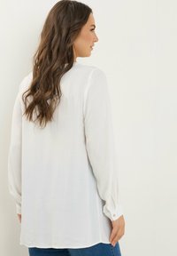 Blusa bianca a maniche lunghe realizzata in tessuto leggero con una vestibilità casual, dotata di polsini con bottoni e orlo arrotondato, abbinata a jeans blu.