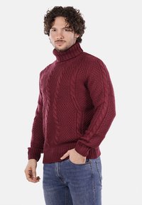 Maglione a collo alto in maglia cable bordeaux, con motivi testurizzati e polsini a costine. Abbinato a jeans di denim.