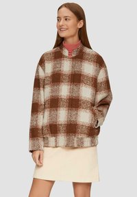 Bruine en witte geruite fleece pullover met een hoge kraag, lange mouwen en vo pockets, gecombineerd met een lichtbeige rok.