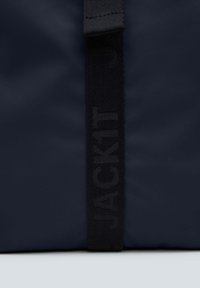 Borsa blu navy in tessuto texture, con una tracolla nera inciisa con la scritta "JACKIT". Il design è elegante e minimalista.