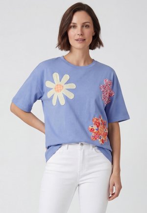 Frau trägt ein blaues kurzärmeliges Hemd mit großen bunten Blumenapplikationen, kombiniert mit weißen Hosen, stehend vor einem schlichten Hintergrund.