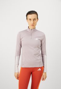 Svijetloplavi sportski gornji dio s polu zatvaračem i dugim rukavima, s bijelim trostrukim naglascima na rukavima i malim logotipom na prsima.