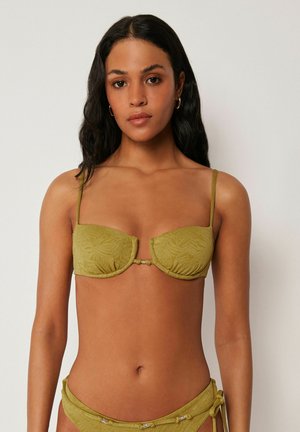 Frau trägt einen strukturierten olivgrünen zweiteiligen Bikini mit goldenen Creolen vor einem neutralen Hintergrund.