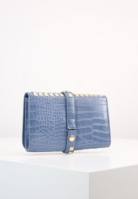 Tyrkysová peněženka s krokodýlovou strukturou, která má klopu se zlatými kovovými akcenty a magnetickým uzávěrem. Kompaktní, obdélníkový design.