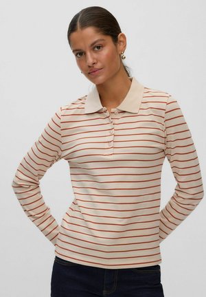 Femme aux cheveux foncés portant des boucles d'oreilles en or et un polo beige à manches longues avec de fines rayures horizontales rouges et un pantalon foncé.