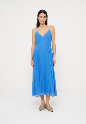 Vrouw met steil donker haar, gekleed in een mouwloze blauwe midijurk met dunne bandjes en beige platte schoenen, staand tegen een witte achtergrond.