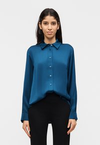 Γυναίκα που φοράει μακρυμάνικο σατέν πουκάμισο σε τυρκουάζ χρώμα με κουμπιά, ελαφρώς μοιρασμένο μέσα σε μαύρο παντελόνι, στέκεται μπροστά σε λευκό απλό φόντο.