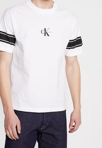 Valkoinen lyhythihainen t-paita, jossa mustat raidat hihassa, ja jossa on pieni logo, joka lukee "CK Calvin Klein Jeans." Kankaana näyttää olevan tasainen puuvilla.