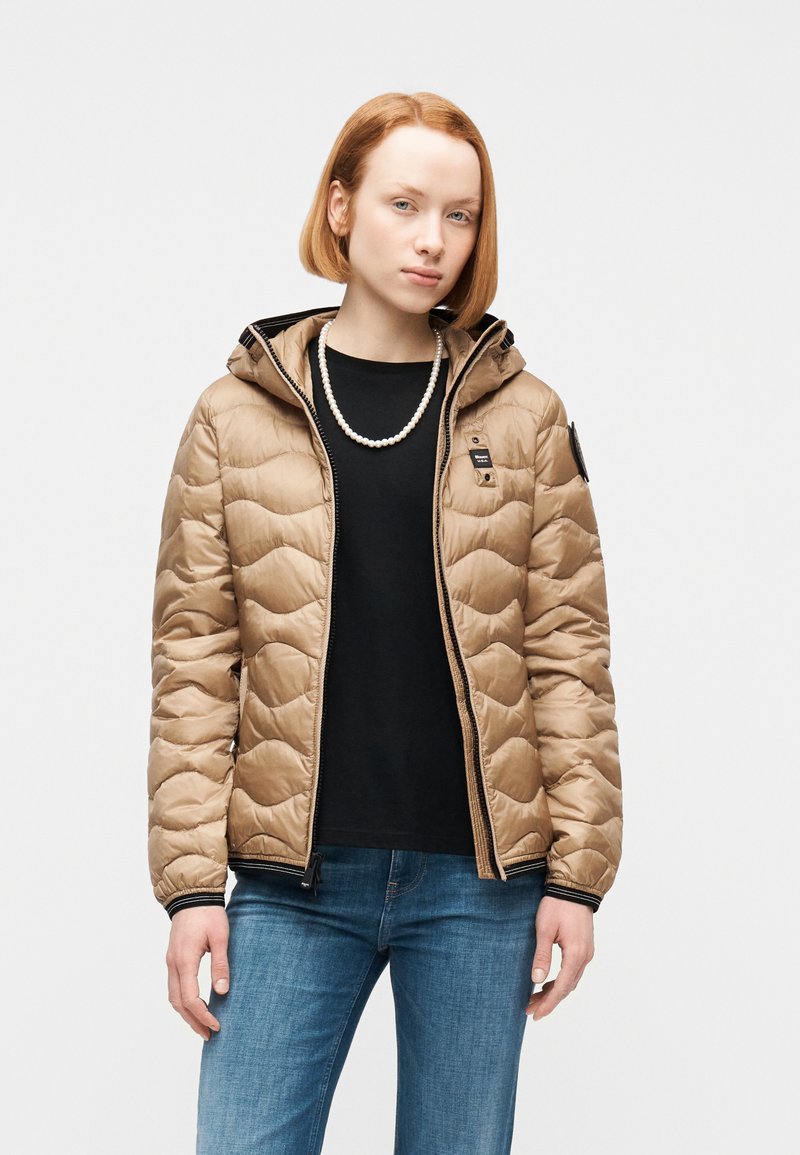 Νεαρή γυναίκα με κοντά κόκκινα μαλλιά φοράει μπεζ καπιτονέ μπουφάν πάνω από μαύρο πουκάμισο, μπλε τζιν και κολιέ με πέρλες, στέκεται μπροστά σε λευκό φόντο.