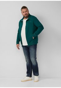 Man met een baard en opgestoken haar in een groene zip-up jas, witte t-shirt, donkere jeans en witte sneakers, staande tegen een grijze muur.