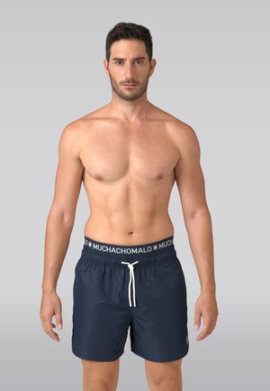 Športovo štíhly muž s tmavými vlasmi, oblečený v tmavomodrých plavkových šortkách s bielou šnúrkou a pásom s nápisom "MUCHACHOMALO", stojaci na sivom jednofarebnom pozadí.