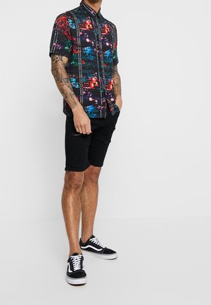 Camicia a maniche corte con bottoni e motivo astratto colorato su sfondo scuro, abbinata a pantaloncini di denim neri e sneakers nere.