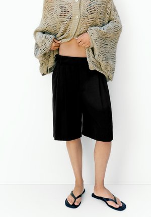 Person hebt beigen gestrickten Pullover hoch, um den Bauch freizulegen, trägt schwarze knielange Shorts und schwarze Flip-Flops, steht vor weißem Hintergrund.
