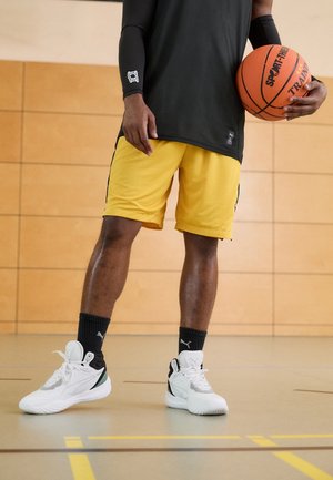 Schwarzes langärmeliges Oberteil, gelbe Basketballshorts aus Mesh mit Seitenstreifen, schwarze Socken und weiße Sportschuhe mit grünen Akzenten. Hält einen Basketball.