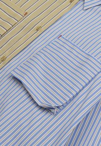 Camicia a righe con sezioni alternate di tessuto khaki e blu, con finitura testurizzata, chiusura a bottoni e taschino sul petto.