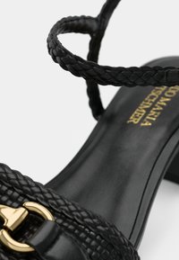Sorte lædersandaler med flettede stropper og en guldbuckle detalje, der har en lav blokhæl og synlig guldbrand tekst på indersålen.