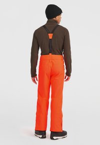 Klarröda snöbyxor med justerbara svarta hängslen, zippered nederdel och rak bendesign, kombinerat med en brun fleece-tröja och svarta stövlar.
