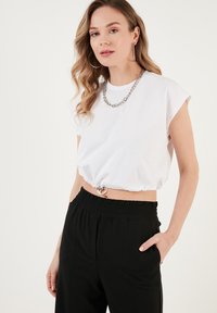 Weiße, kurzärmelige, cropped T-Shirt mit gerafftem Taillenbereich und silbernen Hardwaredetails, kombiniert mit schwarzen Hose mit elastischem Bund.
