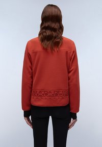Giacca in pile di colore ruggine con logo testurizzato sul retro. Presenta maniche lunghe e una vestibilità rilassata. Tessuto liscio, senza accessori visibili.