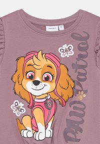 T-shirt di cotone rosa con un cane dei cartoni animati dal pelo marrone, una fascia rosa sulla testa e dettagli a forma di zampa. Maniche arricciate; testo "Paw Patrol" in caratteri scuri.