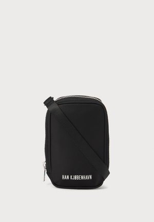 Sac noir de forme rectangulaire, en matériau lisse. Dispose d'une fermeture éclair sur le dessus, d'une bandoulière ajustable, et d'un texte blanc : "HAN KJØBENHAVN."