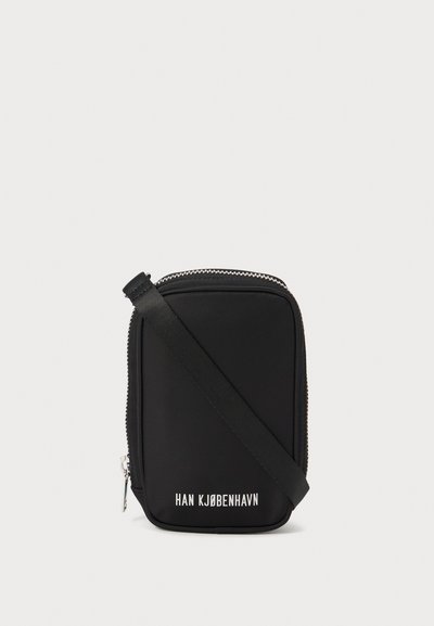 Bolso negro de forma rectangular, hecho de material suave. Cuenta con una cremallera superior, una correa ajustable y texto blanco: "HAN KJØBENHAVN."